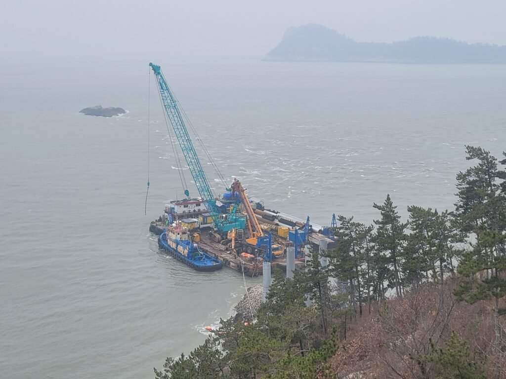 [Photo ③] Site overview. Barge, crawler crane and tug working against the rock shoreline, with land-section piles already standing.