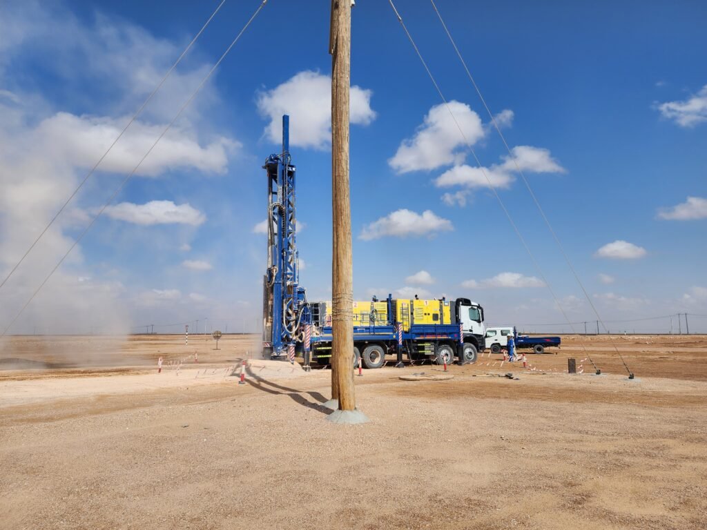 Truck-mounted drill rig drilling a Ø1,000 mm pole hole beside an erected pole on an oilfield overhead line